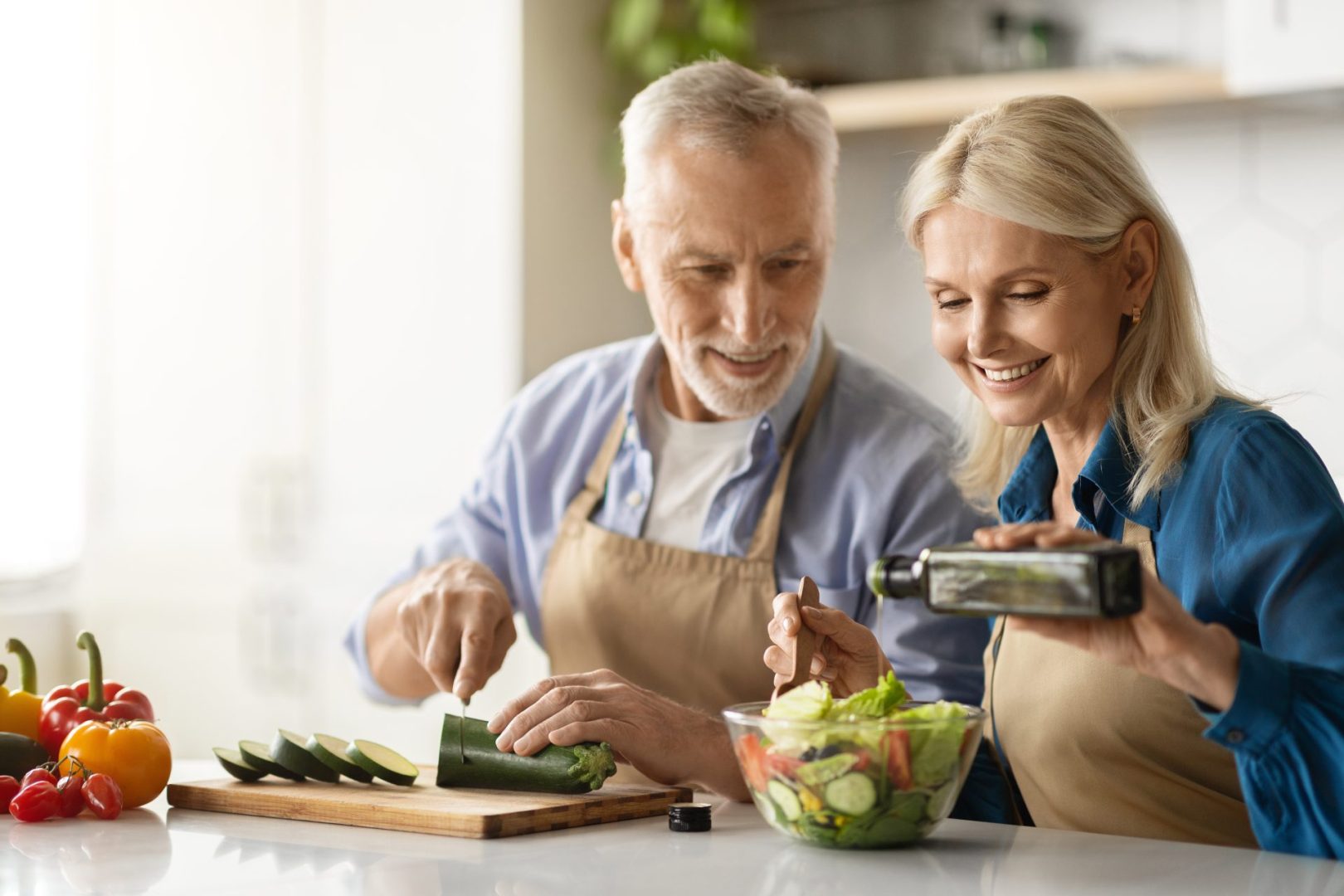 Happy Senior Couple Preparing Tasty Lunch Together In Kitchen, Smiling Mature Man And Woman Cooking Healthy Vegetarian Meal At Home, Woman Adding Oil To Fresh Vegetable Salad, Free Space